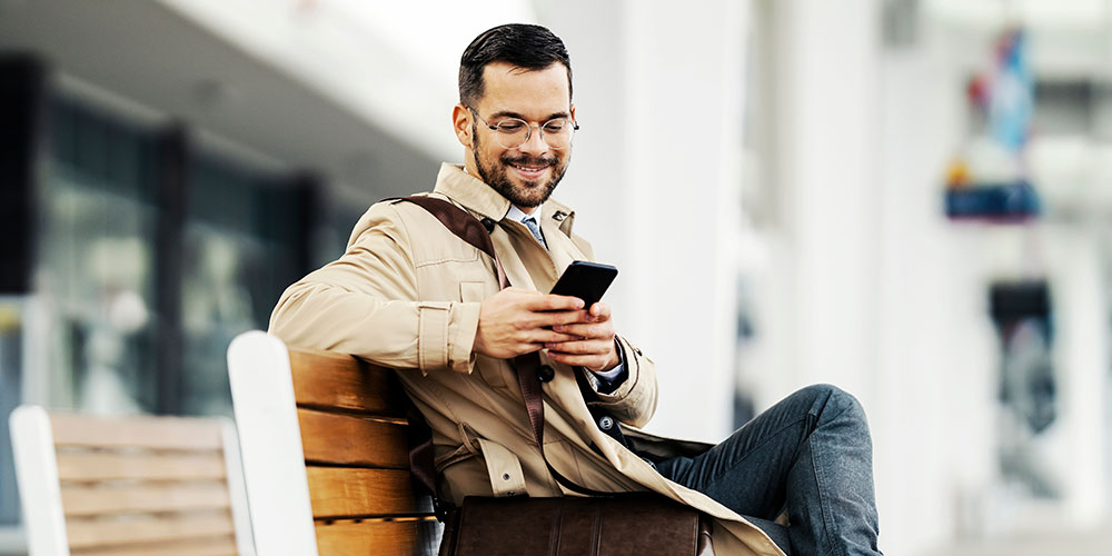 Ein Mann sitzt am Bahnhof auf einer Bank und schaut lächelnd auf sein Handy.