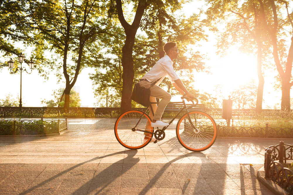 Mann fährt auf einem Fahrrad eine Promenade entlang, im Hintergrund scheint die untergehende Sonne durch Bäume.