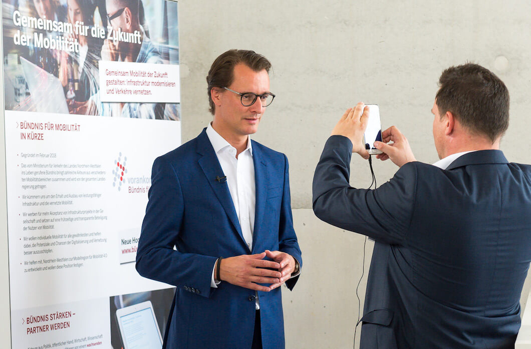 Zwei Männer in Anzügen stehen vor einem Pappaufsteller, der eine fotografiert den anderen mit einem Smartphone.