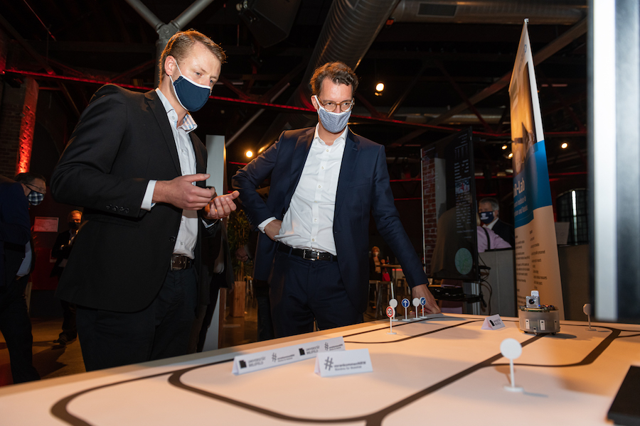 Zwei Männer in Hemd und Anzug mit Mund-Nase-Bedeckung stehen an einem Tisch, auf dem ein Verkehrsmodell aufgebaut ist.