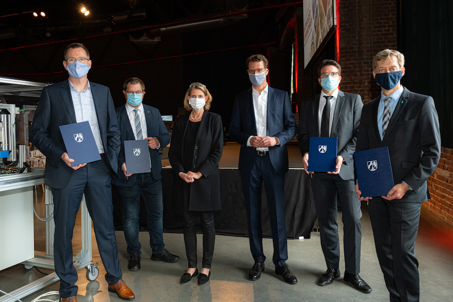 Fünf Männer und eine Frau in Anzug, Blazer stehen nebeneinander mit Maske und halten Dokumente in der Hand, auf denen das NRW-Logo ist.